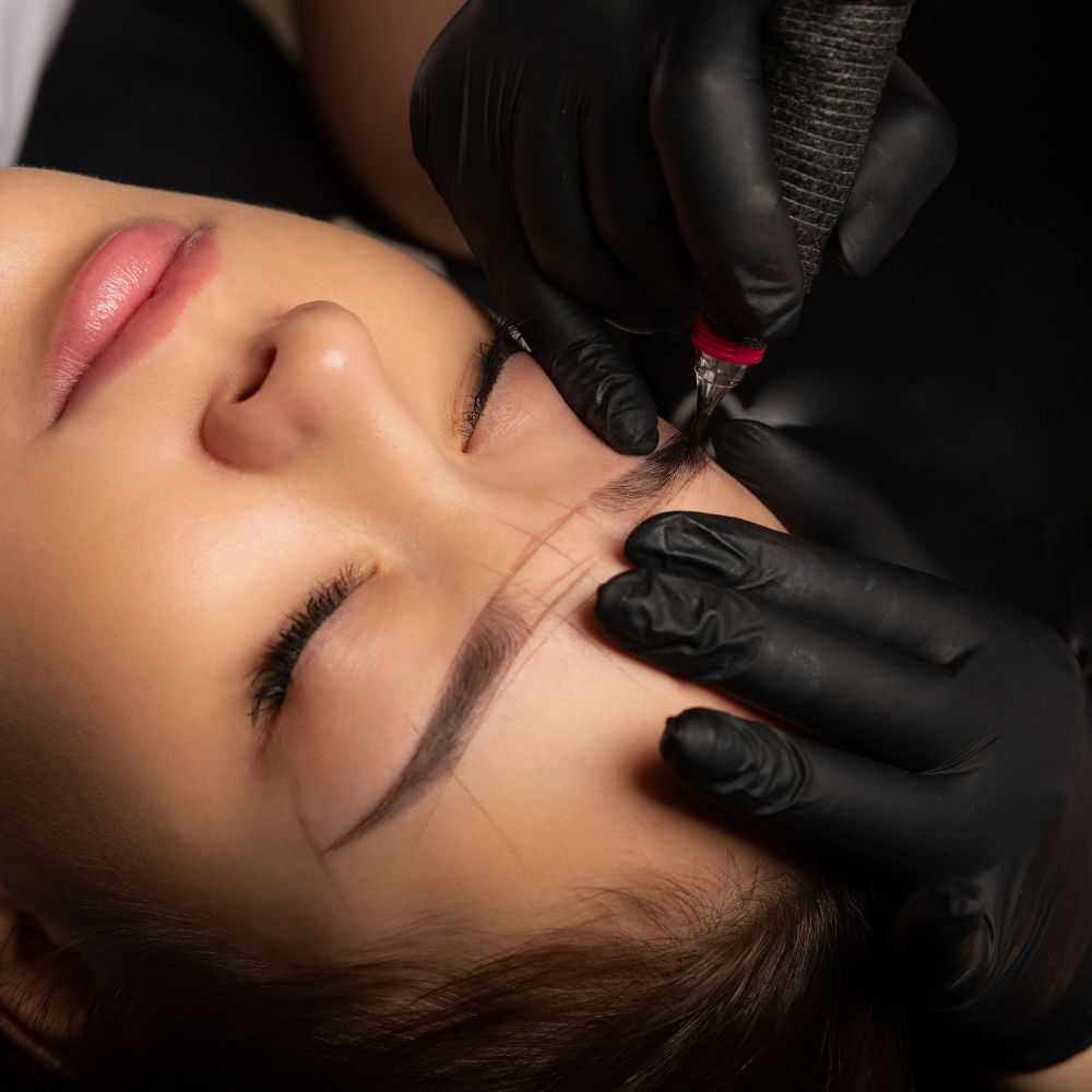 Kobieta leży z zamkniętymi oczami podczas zabiegu makijażu permanentnego brwi. Specjalista w czarnych rękawiczkach wykonuje pigmentację skóry przy użyciu urządzenia do mikropigmentacji.